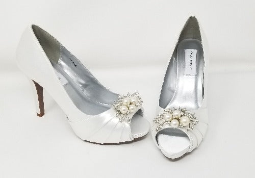 A pair of high heeled white satin shoes with a peep toe and a hidden platform at the front of the shoes and a crystal design on the front of the shoes